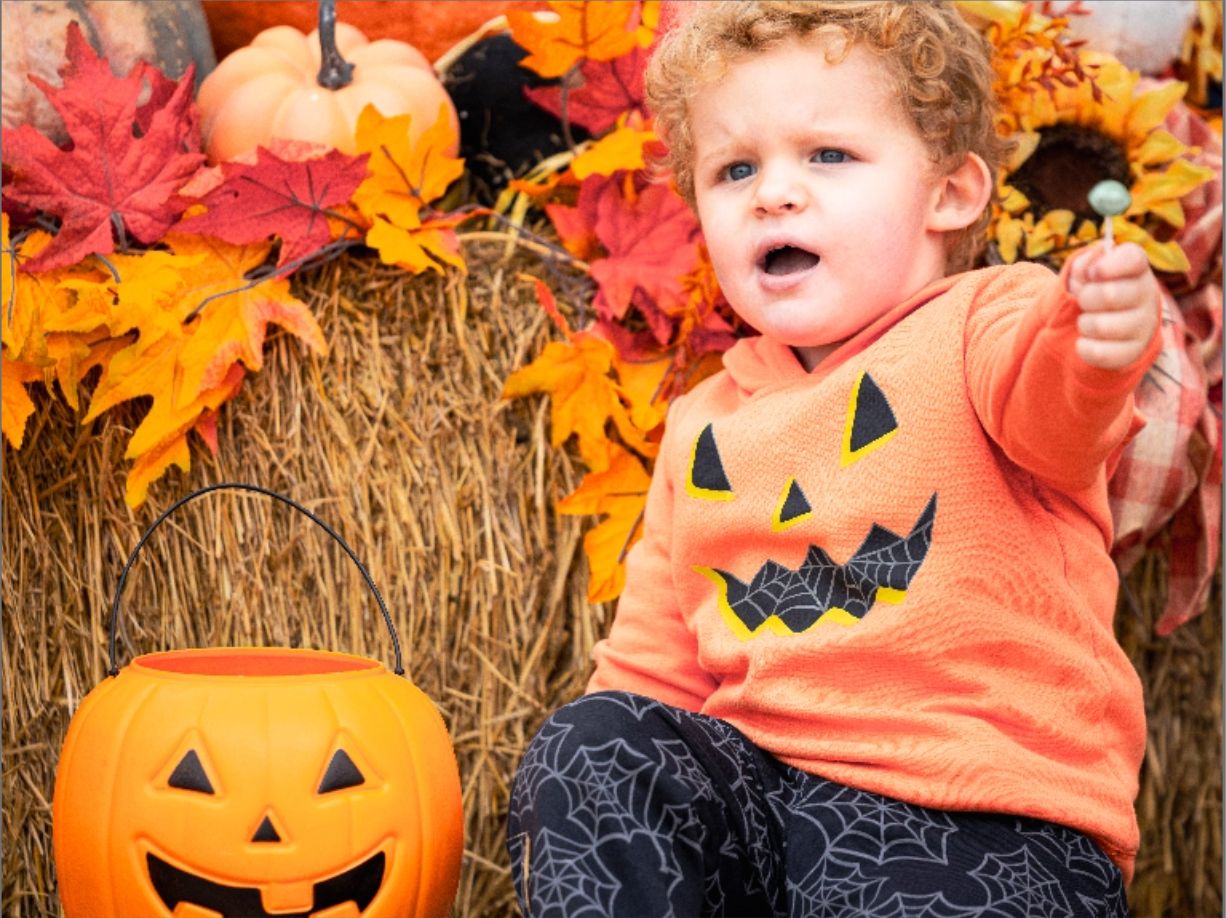 A child at Trick or Treat Main Street wearing a jack-o-lantern hoodie and spider web pants seated on a bench next to a jack-o-lantern, and in the background, a bale of hay with pumpkins and leaves displayed on top.